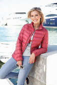 A blonde woman wearing jeans, a top and quilted jacket standing at a harbour