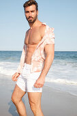 Junger Mann mit offenem Hemd und Shorts steht am Strand