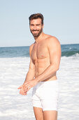 Junger Mann mit Shorts und nacktem Oberkörper steht am Strand