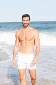 A young topless man walking along a beach wearing shorts