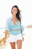 A brunette woman on a beach wearing a light-blue bathing suit, a shirt and white shorts