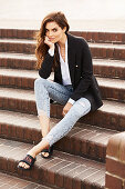 A brunette woman wearing a white shirt, a black blazer and jeans sitting on steps