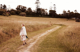 A blonde woman on a field path wearing a floral patterned dress and a light jacket