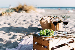 Two glasses with red wine and plate with white grape standing on timber box on sandy beach.
