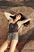 A brunette woman wearing a brown top, a jacket and shorts lying on rocks