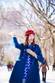 A brunette woman wearing a red hat and a blue coat throwing a snowball in a wintery park