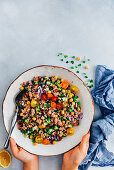 Hands holding a bowl of tuna salad with chickpeas, peas, tomatoes, cucumbers, onion and herbs