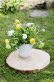 Spring bouquet of tulips, ranunculus and ox-eye daisies in jug on slice of tree trunk