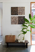 Basket, candle lantern and leafy branch on bench below two-tone artworks on wall