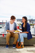 Junges Pärchen sitzt auf einer Bank und teilt sich Fast Food Essen