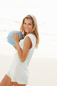 Reife blonde Frau mit blauem Ball in weißem Longshirt am Strand