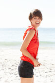 A mature brunette woman on a beach wearing a red shirt, a necklace and black shorts