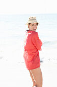 A mature brunette woman on a beach wearing a red shirt and a beige hat