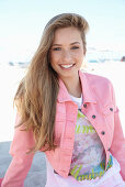 A young blonde woman on a beach wearing a printed t-shirt and a pink denim jacket