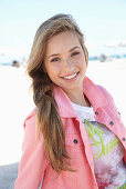 A young blonde woman on a beach wearing a printed t-shirt and a pink denim jacket