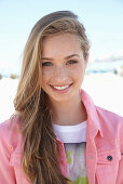 A young blonde woman on a beach wearing a t-shirt and a pink denim jacket