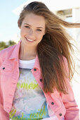 A young blonde woman on a beach wearing a printed t-shirt and a pink denim jacket