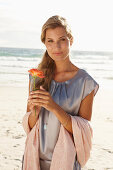 Reife blonde Frau mit silbernem Sommerkleid und orangener Blume am Strand