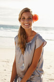 A mature blonde woman on a beach wearing a silver summer dress with an orange flower behind her ear