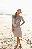 A mature blonde woman on a beach wearing a silver summer dress with an orange flower behind her ear