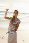 A mature blonde woman on a beach wearing a silver summer dress and holding a white balloon