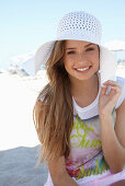 A young blonde woman on a beach wearing a colourful t-shirt and a white summer hat