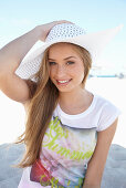 A young blonde woman on a beach wearing a colourful t-shirt and a white summer hat