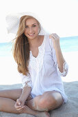 A young blonde woman on a beach wearing a white summer dress and a white summer hat