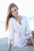 A young blonde woman on a beach wearing a white summer dress and a pink necklace