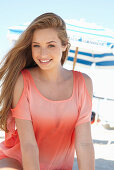 A young blonde woman on a beach wearing a salmon-coloured, off-the-shoulder top