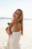 A mature blonde woman on a beach wearing lingerie and a cardigan and holding a flower