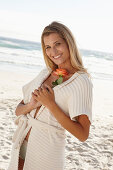 A mature blonde woman on a beach wearing lingerie and a cardigan and holding a flower