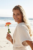 A mature blonde woman on a beach wearing lingerie and a cardigan and holding a flower