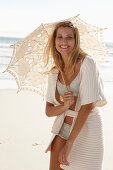 A mature blonde woman on a beach wearing lingerie and a cardigan with a parasol