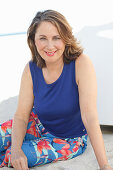 A brunette woman on a beach wearing a purple top and colourful trousers