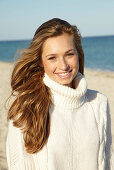 A young blonde woman on a beach wearing a white turtle neck jumper