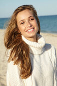 A young blonde woman on a beach wearing a white turtle neck jumper