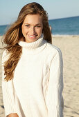 A young blonde woman on a beach wearing a white turtle neck jumper