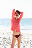 A mature brunette woman on a beach wearing a black bikini and a red shirt