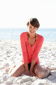 A mature brunette woman on a beach wearing a black bikini and a red shirt