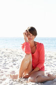 A mature brunette woman on a beach wearing a black bikini and a red shirt