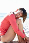 A mature brunette woman on a beach wearing a black bikini and a red shirt