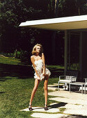 A young blonde woman posing in the garden of a house with a terrace wearing a beige evening dress