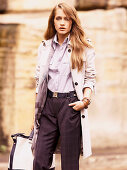 A young woman wearing purple trousers, a blouse and a trench coat with a bag