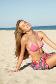 A young blonde woman by the sea wearing a pink bikini with a beach towel around her hips