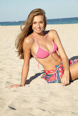 A young blonde woman by the sea wearing a pink bikini with a beach towel around her hips