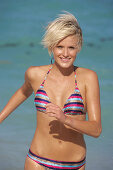 A mature blonde woman with short hair on a beach wearing a striped bikini