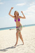 A young blonde woman on a beach wearing a purple bikini with a towel around her hips