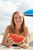 A young blonde woman on a beach wearing a colourful summer dress holding a wedge of melon