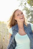 A young blonde woman outside wearing a sleeveless denim jacket and a light-blue top